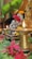 A traditional spiritual setup features a small statue holding bright red flowers and surrounded by lit brass oil lamps. The arrangement includes vibrant pink flowers and large green leaves, creating a serene and devotional atmosphere.