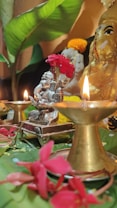 A traditional spiritual setup features a small statue holding bright red flowers and surrounded by lit brass oil lamps. The arrangement includes vibrant pink flowers and large green leaves, creating a serene and devotional atmosphere.