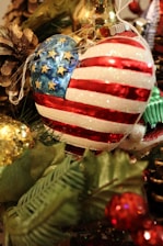 A heart-shaped ornament decorated with glittering stars and stripes resembling the American flag is nestled among green pine branches and other Christmas decorations. Nearby are a pine cone and gold bauble, adding to the festive atmosphere.
