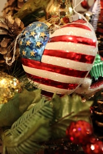 A heart-shaped ornament decorated with glittering stars and stripes resembling the American flag is nestled among green pine branches and other Christmas decorations. Nearby are a pine cone and gold bauble, adding to the festive atmosphere.