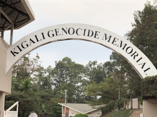 An arched sign displaying the words 'Kigali Genocide Memorial' stands prominently in the foreground. It is surrounded by lush greenery, with various trees and shrubs visible. In the background, part of a building with a light-colored roof can be seen, along with more trees and a cloudy sky.