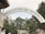 An arched sign displaying the words 'Kigali Genocide Memorial' stands prominently in the foreground. It is surrounded by lush greenery, with various trees and shrubs visible. In the background, part of a building with a light-colored roof can be seen, along with more trees and a cloudy sky.