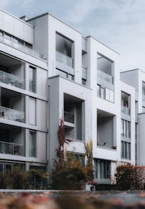 A modern, multi-story apartment building with large balconies and a minimalist architectural design. The structure features sharp lines, geometric shapes, and a facade dominated by white and gray tones. Climbing plants with autumn-colored leaves add a touch of nature to the urban environment. The sky is overcast, contributing to a calm and neutral ambience.