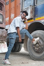 A person is posing confidently with one foot on the tire of a large truck. Wearing sunglasses, a light blue striped shirt, and blue jeans, the individual is casually leaning against the vehicle. The truck has an orange and blue color scheme with some text written on the side.