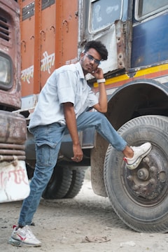 A person is posing confidently with one foot on the tire of a large truck. Wearing sunglasses, a light blue striped shirt, and blue jeans, the individual is casually leaning against the vehicle. The truck has an orange and blue color scheme with some text written on the side.