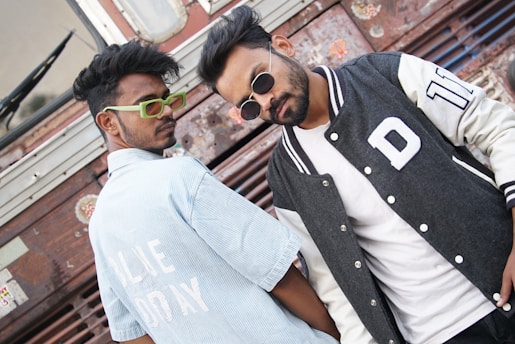 Two individuals are posing in front of a textured metal background, showcasing a casual and stylish demeanor. One is wearing a light blue striped shirt with bold white text, along with vibrant green glasses. The other person sports a black and white varsity jacket layered over a plain white shirt and wears dark sunglasses. Their expressions are confident and relaxed.