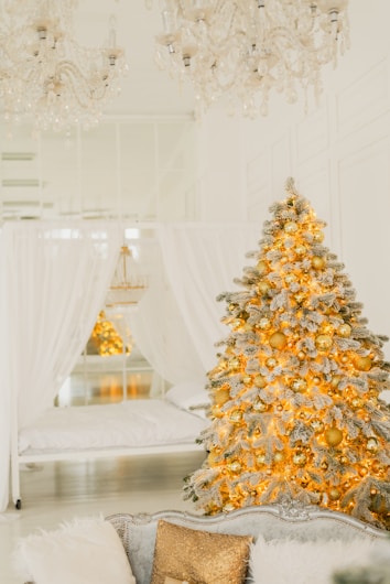 A beautifully decorated Christmas tree adorned with golden ornaments and glowing lights stands in a luxurious, softly-lit white room. Elegant chandeliers hang from the ceiling, adding to the opulent ambiance. White drapes surround a bed in the background, contributing to the room's serene and festive atmosphere.