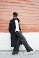 A man dressed in a classic grey coat and light blue shirt, leaning casually against a stone wall with a thoughtful gaze.