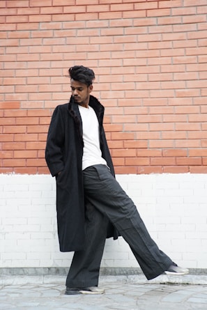 A man dressed in a classic grey coat and light blue shirt, leaning casually against a stone wall with a thoughtful gaze.
