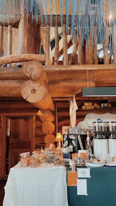 A cozy, wooden interior with natural log beams and a rustic aesthetic. A small setup of what appears to be a coffee or snack station with various items displayed on tables covered with white cloth. There are bags, jars, and a coffee grinder or espresso machine present. A warm lamp adds a touch of light to the scene.