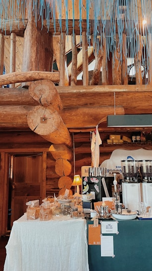 A cozy, wooden interior with natural log beams and a rustic aesthetic. A small setup of what appears to be a coffee or snack station with various items displayed on tables covered with white cloth. There are bags, jars, and a coffee grinder or espresso machine present. A warm lamp adds a touch of light to the scene.