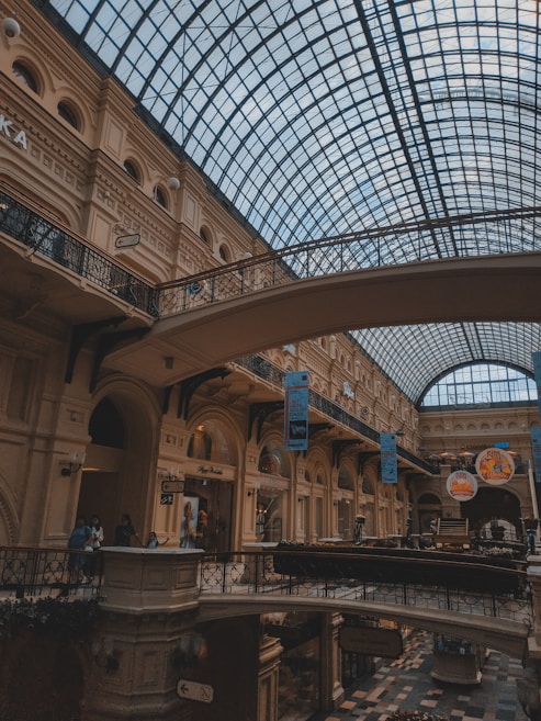 A spacious, elegantly designed indoor mall with a high arched glass ceiling allowing natural light to illuminate the space. The architecture features classical elements with ornately decorated arches and balconies. Shoppers can be seen walking along the upper levels, and various store signs and advertisements are visible.