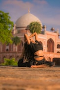 A person is performing a yoga pose in the foreground, with an ornate historic building featuring a large dome in the background. The setting appears to be outdoors, with lush green foliage and a clear blue sky adding to the serene atmosphere.