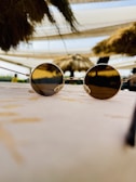 A pair of round sunglasses with dark lenses is placed on a textured surface, possibly a table. The background features out-of-focus thatched roofing and a bright sky, suggesting an outdoor setting.