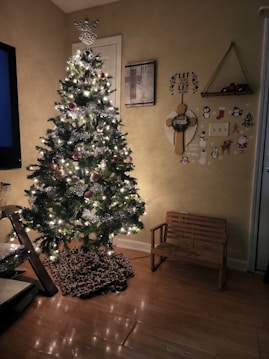 A decorated Christmas tree stands in a cozy living room, adorned with lights, baubles, and a silver garland. The floor has a soft animal-print tree skirt, and a small wooden bench is positioned nearby. Wall decorations include a motivational plaque, a Christmas-themed wreath, and festive stickers featuring penguins and Santa Claus. Warm lighting enhances the holiday atmosphere.