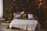 A cozy bedroom setup with a neatly made bed adorned with various pillows in shades of gray and mustard yellow. The room is warmly lit by string lights that hang above the bed, and a small lit Christmas tree stands in the corner with a few wrapped gifts beneath it. There are decorative shelves with plants and ornaments along the wall and a soft, textured rug on the floor.