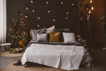 A cozy bedroom setup with a neatly made bed adorned with various pillows in shades of gray and mustard yellow. The room is warmly lit by string lights that hang above the bed, and a small lit Christmas tree stands in the corner with a few wrapped gifts beneath it. There are decorative shelves with plants and ornaments along the wall and a soft, textured rug on the floor.