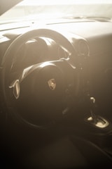 A car interior with a focus on the steering wheel, which features an emblem at its center. The dashboard and part of a gear shift can also be seen. The scene is bathed in soft, ambient lighting, creating a warm and somewhat ethereal atmosphere.