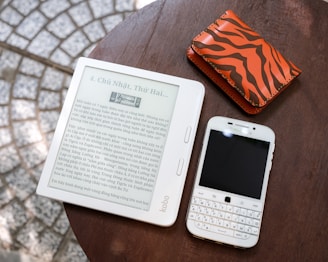 A Kobo e-reader displaying text in a foreign language, placed on a wooden surface next to a white Blackberry smartphone with a physical keyboard and an orange wallet with a brown animal-print pattern.