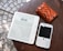 A Kobo e-reader displaying text in a foreign language, placed on a wooden surface next to a white Blackberry smartphone with a physical keyboard and an orange wallet with a brown animal-print pattern.