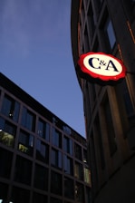 A cityscape view at twilight featuring modern office buildings with large glass windows reflecting the dimming sky. One building prominently displays a bright illuminated sign with 'C&A' on it, emphasizing its presence against the darker surroundings.