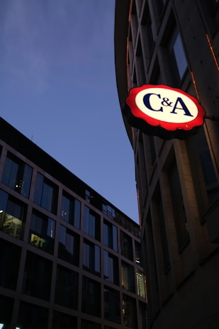 A cityscape view at twilight featuring modern office buildings with large glass windows reflecting the dimming sky. One building prominently displays a bright illuminated sign with 'C&A' on it, emphasizing its presence against the darker surroundings.