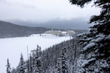 A snowy landscape with dense pine forests surrounding a frozen lake. In the background, a large hotel complex sits against the backdrop of distant, snow-capped mountains under a cloudy sky. Snowy branches are visible in the foreground.