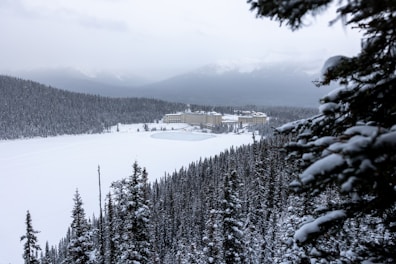 A snowy landscape with dense pine forests surrounding a frozen lake. In the background, a large hotel complex sits against the backdrop of distant, snow-capped mountains under a cloudy sky. Snowy branches are visible in the foreground.