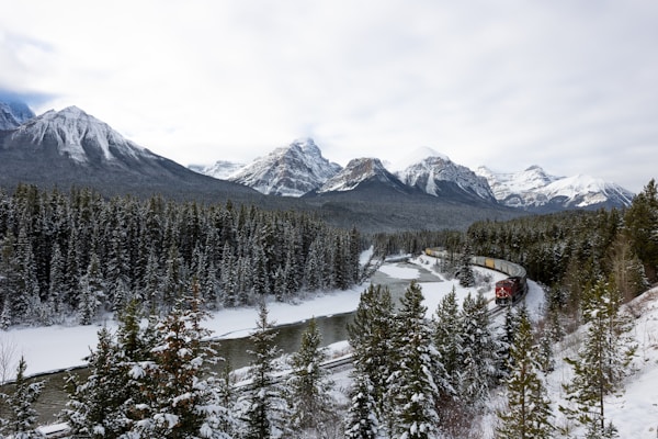 A scenic landscape features snow-covered mountains under an overcast sky, with lush, evergreen trees blanketing the foothills. A river winds through the forest, and a red train is seen curving along the tracks, adding a dynamic element to the serene environment.
