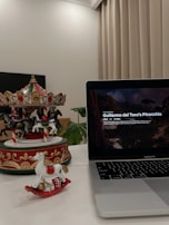 A decorative carousel and a small rocking horse ornament are placed on a white surface. The carousel features intricate designs with horses, and the rocking horse is white with a red base. A MacBook Pro displaying the title 'Guillermo del Toro's Pinocchio' occupies the right side of the image. The background includes a green plant and beige curtains.