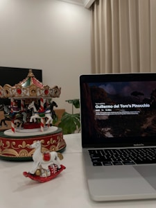 A decorative carousel and a small rocking horse ornament are placed on a white surface. The carousel features intricate designs with horses, and the rocking horse is white with a red base. A MacBook Pro displaying the title 'Guillermo del Toro's Pinocchio' occupies the right side of the image. The background includes a green plant and beige curtains.