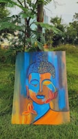 A colorful painting featuring an abstract representation of a serene face in shades of blue, orange, and yellow. The artwork is adorned with lotus flowers and placed against a backdrop of lush green grass and trees.