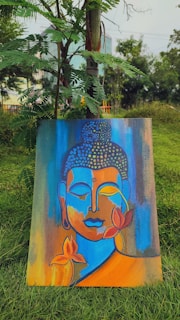 A colorful painting featuring an abstract representation of a serene face in shades of blue, orange, and yellow. The artwork is adorned with lotus flowers and placed against a backdrop of lush green grass and trees.