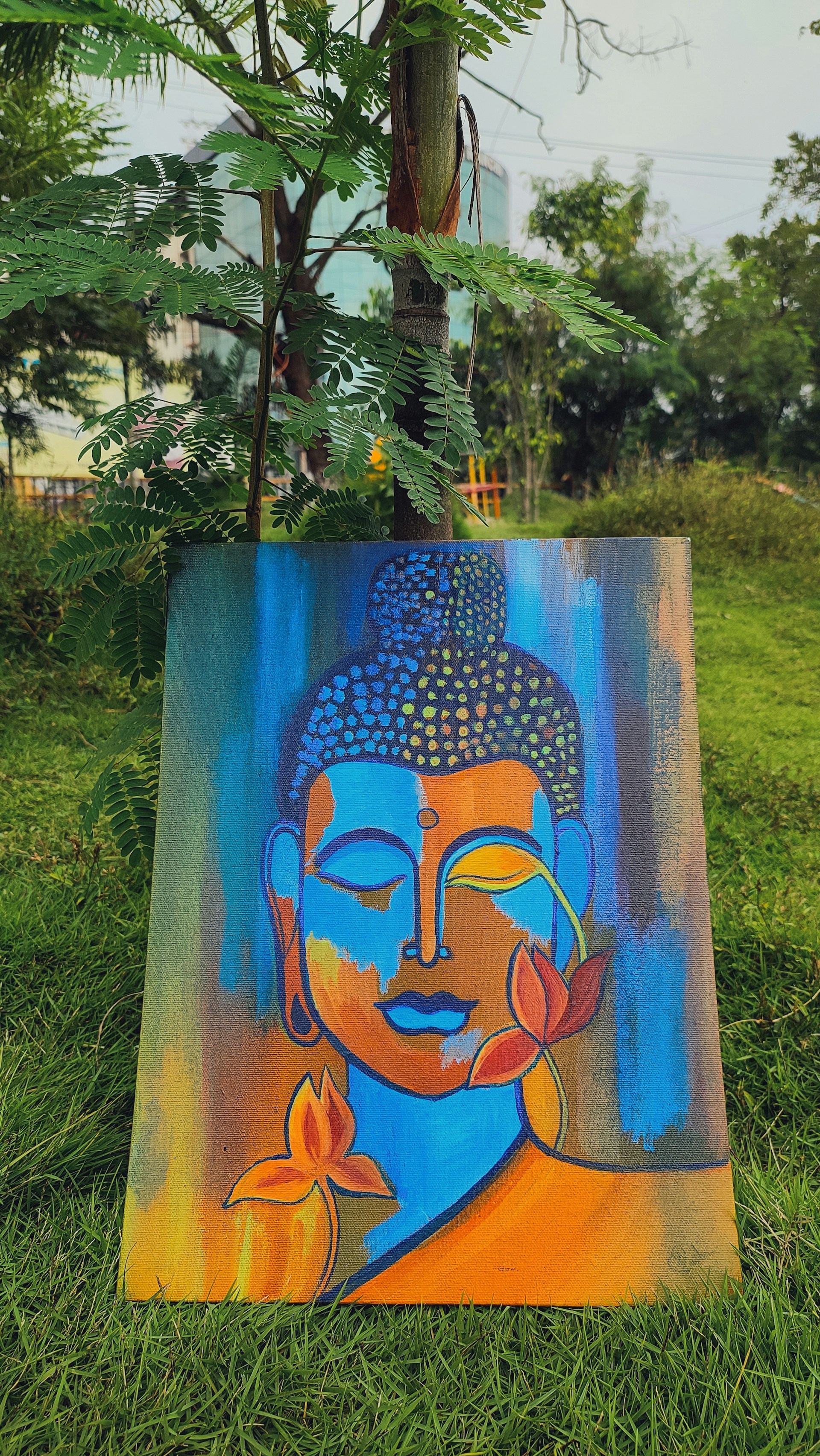 A colorful painting featuring an abstract representation of a serene face in shades of blue, orange, and yellow. The artwork is adorned with lotus flowers and placed against a backdrop of lush green grass and trees.