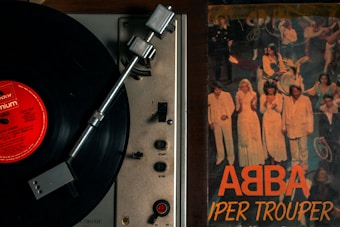 A vintage record player with a black vinyl record spinning, placed next to an album cover. The album cover features a group of people on stage, wearing coordinated outfits in white and light colors. The title on the album cover reads 'ABBA Super Trouper' in bold orange letters.