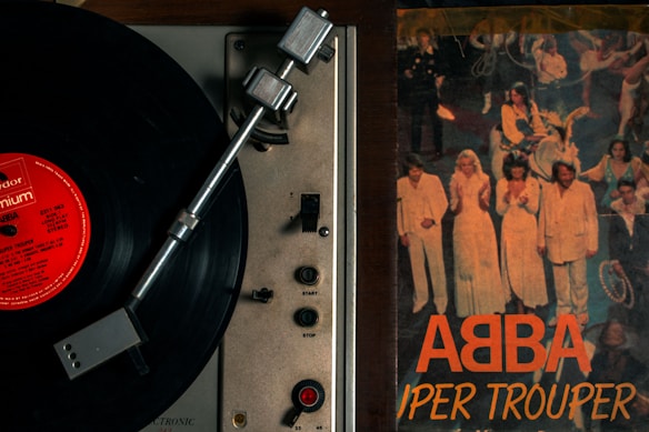 A vintage record player with a black vinyl record spinning, placed next to an album cover. The album cover features a group of people on stage, wearing coordinated outfits in white and light colors. The title on the album cover reads 'ABBA Super Trouper' in bold orange letters.