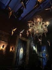 A room with a decorative, wire-framed chandelier hangs from a high ceiling. Golden star-shaped ornaments are suspended around it. Brick walls adorned with paintings and modern wall sconces add character. There's a cozy and artistic atmosphere.