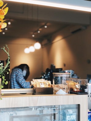 A cozy cafe interior with a warm ambiance. In the foreground, a counter displays a variety of packaged snacks and baked goods, alongside decorative items. The background is softly lit, with a person seated, wearing a striped sweater and a face mask.