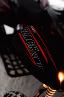 A close-up of a snowmobile featuring a part with the text 'BLACK EDITION' written on it. The snowmobile has a sleek design with a combination of black and red colors. Part of the suspension and a wheel on snow are visible. A light on the vehicle is shining brightly.