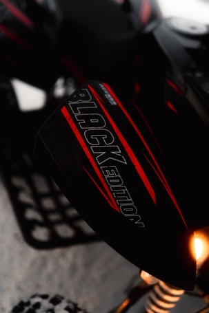 A close-up of a snowmobile featuring a part with the text 'BLACK EDITION' written on it. The snowmobile has a sleek design with a combination of black and red colors. Part of the suspension and a wheel on snow are visible. A light on the vehicle is shining brightly.