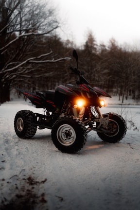 A four-wheeled all-terrain vehicle with headlights on, positioned on a snowy path in a forest setting. The surrounding trees are covered with a light layer of snow, creating a wintery atmosphere.