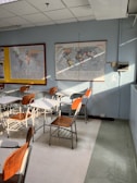 A classroom setting with several empty wooden and metal chairs arranged around desks. Wall-mounted maps are visible, with one world map displaying various countries. Sunlight streams in, casting shadows on the floor.