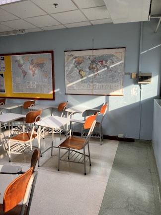 A classroom setting with several empty wooden and metal chairs arranged around desks. Wall-mounted maps are visible, with one world map displaying various countries. Sunlight streams in, casting shadows on the floor.