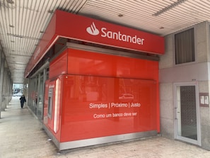 A red bank branch exterior with the Santander logo prominently displayed. The building features large windows and a translated slogan that emphasizes simplicity and fairness. A person is walking on the sidewalk next to the building.