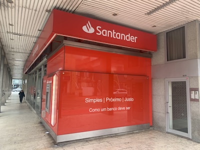 A red bank branch exterior with the Santander logo prominently displayed. The building features large windows and a translated slogan that emphasizes simplicity and fairness. A person is walking on the sidewalk next to the building.