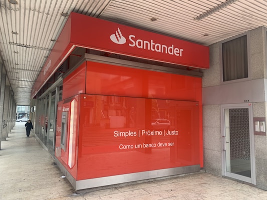 A red bank branch exterior with the Santander logo prominently displayed. The building features large windows and a translated slogan that emphasizes simplicity and fairness. A person is walking on the sidewalk next to the building.