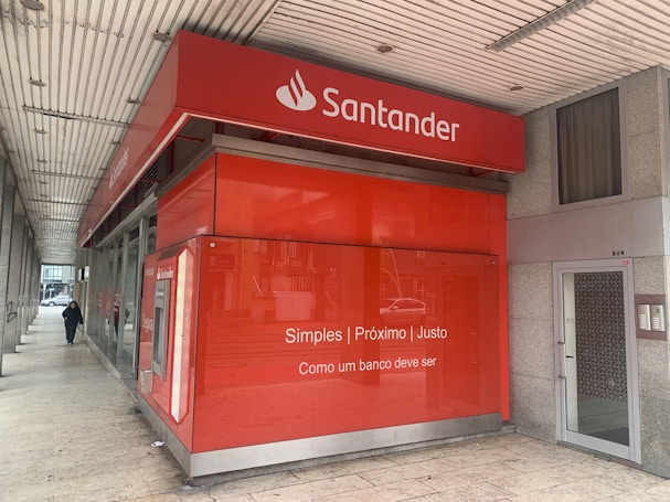 A red bank branch exterior with the Santander logo prominently displayed. The building features large windows and a translated slogan that emphasizes simplicity and fairness. A person is walking on the sidewalk next to the building.