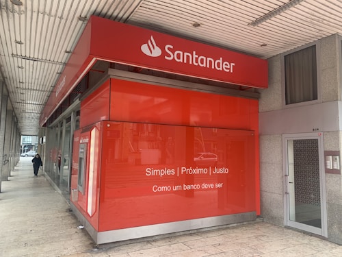 A red bank branch exterior with the Santander logo prominently displayed. The building features large windows and a translated slogan that emphasizes simplicity and fairness. A person is walking on the sidewalk next to the building.