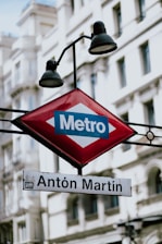 A metro station sign with the name 'Antón Martín' prominently displayed beneath a red diamond-shaped Metro emblem. The background features a classical building with arched windows.