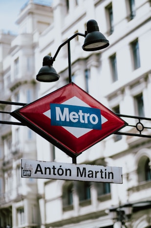 A metro station sign with the name 'Antón Martín' prominently displayed beneath a red diamond-shaped Metro emblem. The background features a classical building with arched windows.
